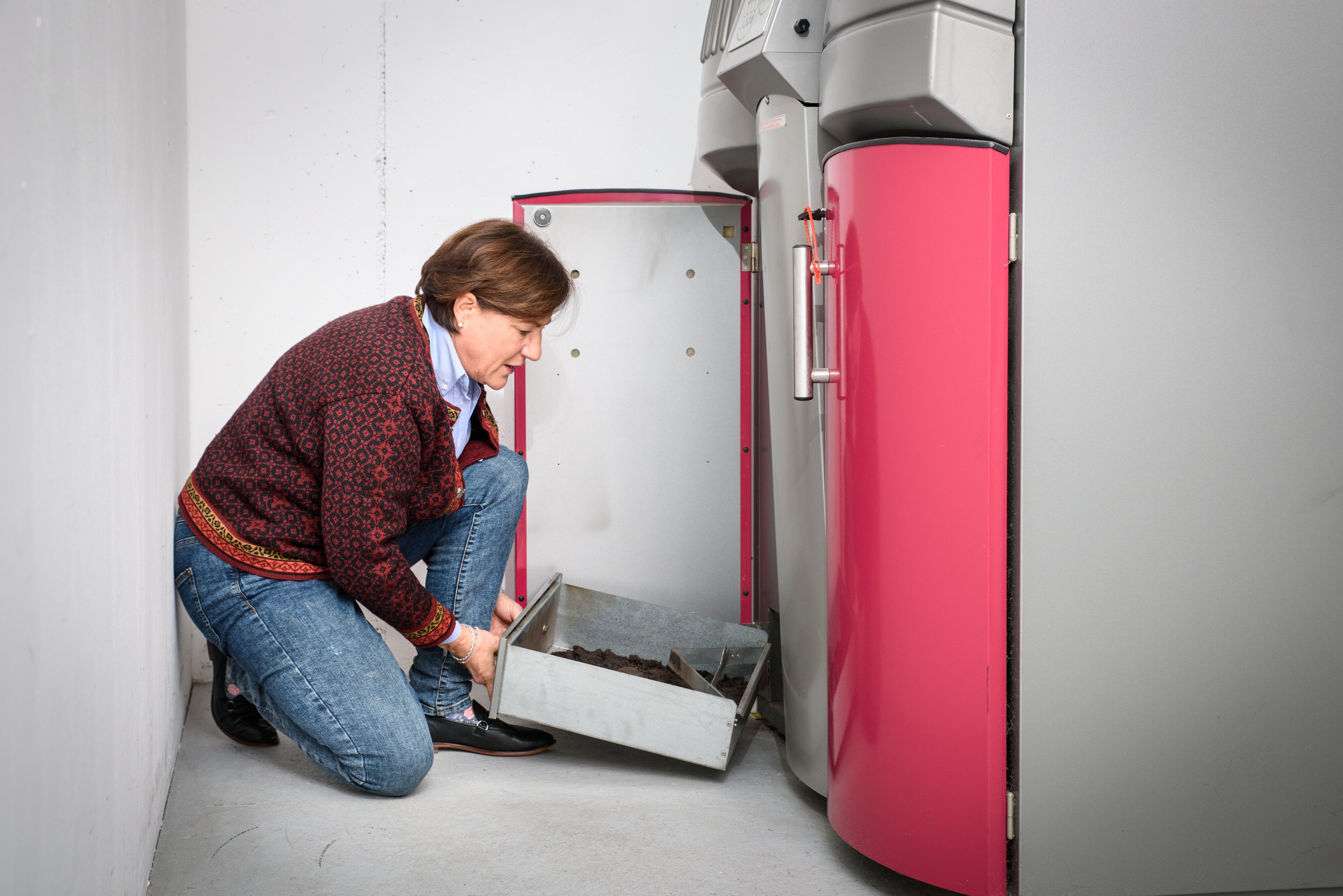 Frau mittleren Alters kniet links auf dem Boden, rechts von ihr ein moderner Pelletheizkessel in grau und rot. Sie hält den Aschekasten in den Händen.