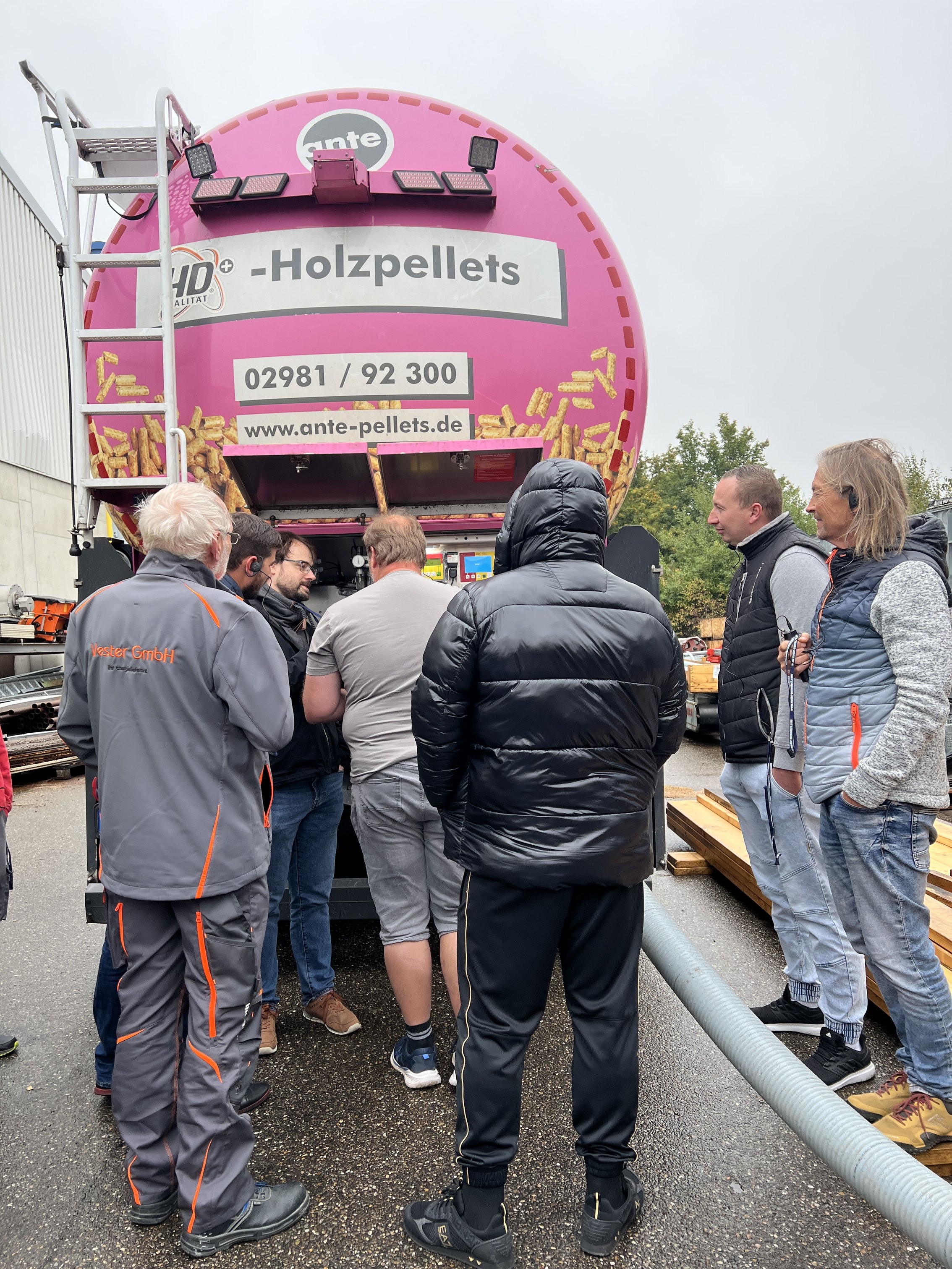 Das Bild im Hochformat zeigt einen pinken Lkw mit Pellets. Davor eine Gruppe Menschen im Gespräch.