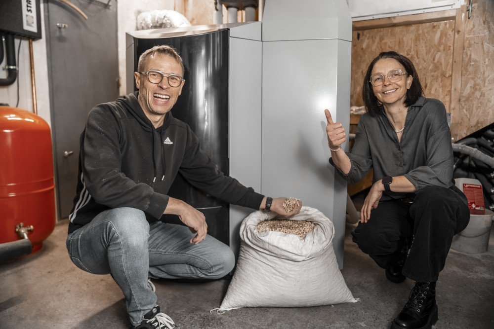 Das Bild zeigt zwei Personen in einem Kellerraum vor einem Sack gefüllt mit Pellets. Die männliche Person (links) hält in einer Hand Pellets. Dahinter ist ein Pelletkessel zu sehen.